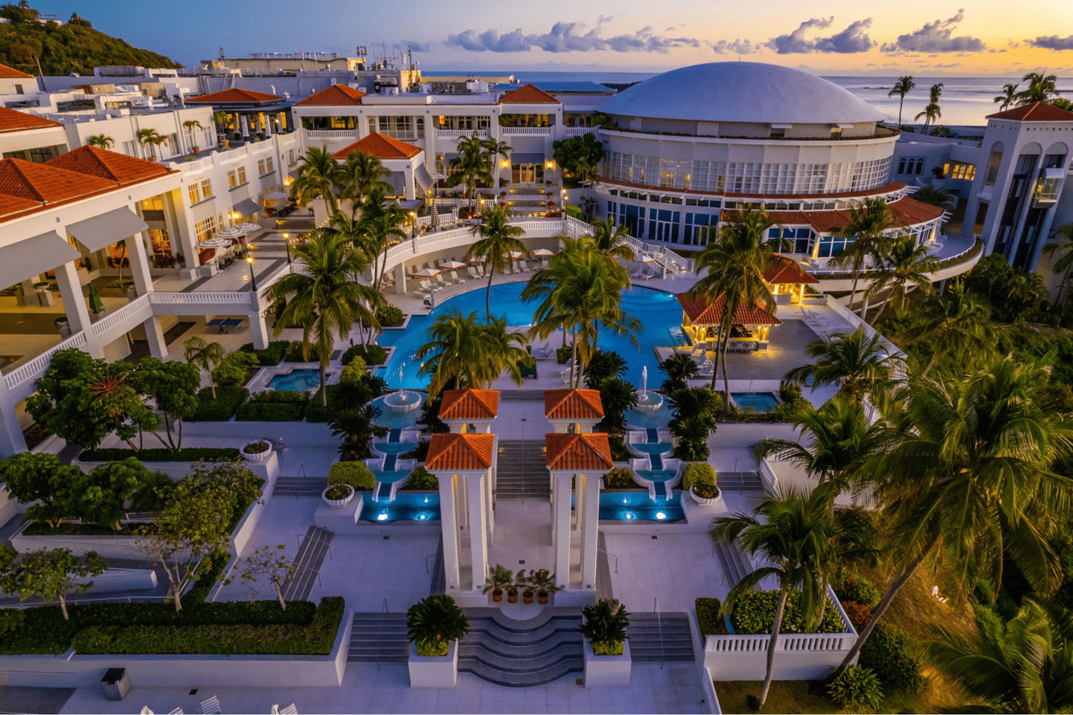 A luxurious resort with a pool, palm trees, and ocean view at sunset.