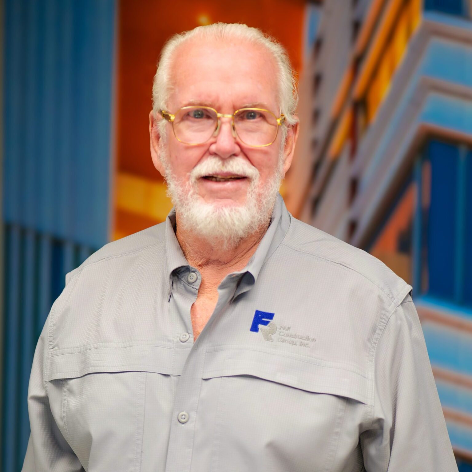 Elderly man with white beard and glasses smiles, wearing a gray shirt with a logo.