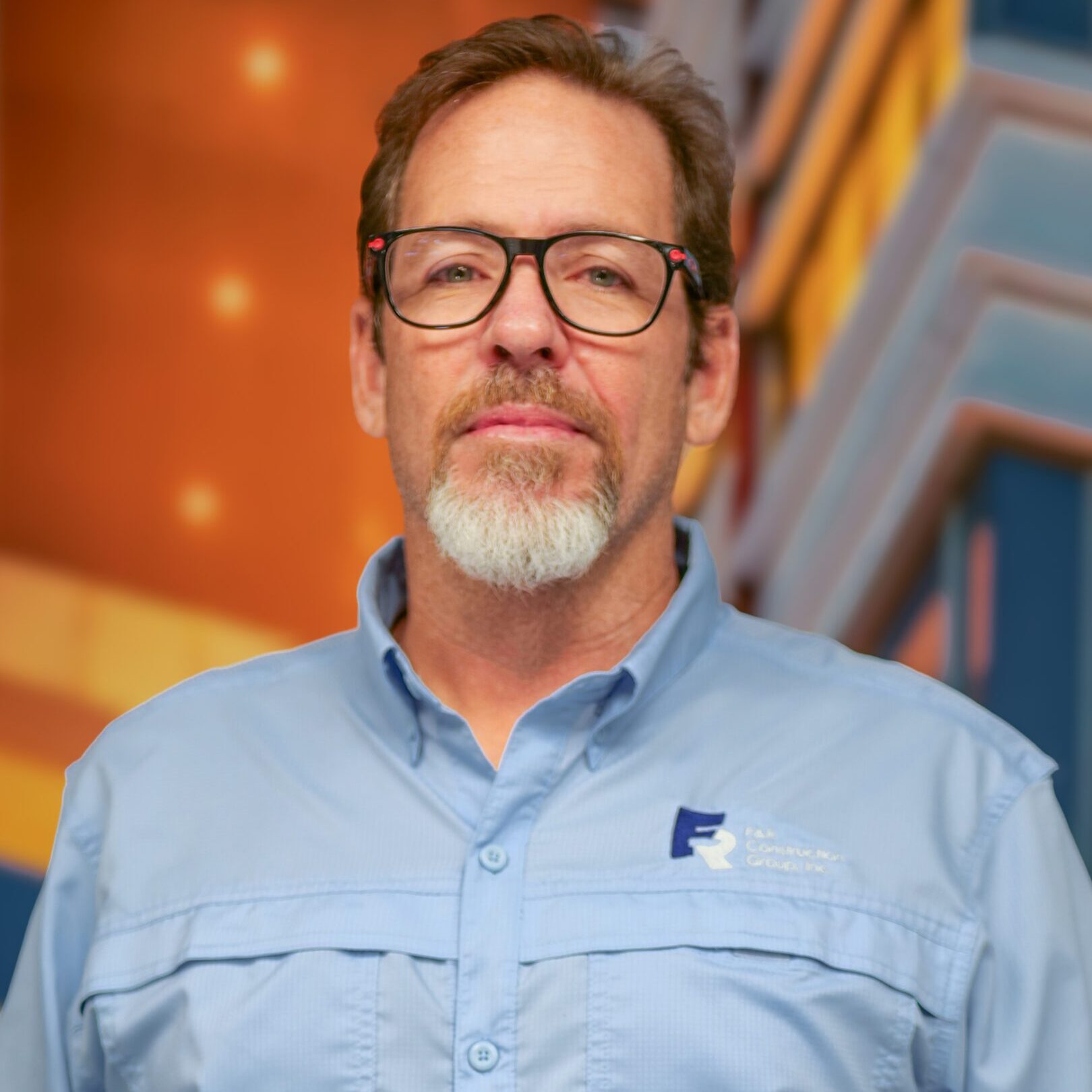 Middle-aged man with glasses and beard, wearing a light blue shirt indoors.